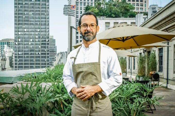 Um homem posa de avental no terraço de um restaurante. Há prédios no fundo e dá para ver a bandeira de São Paulo em um deles