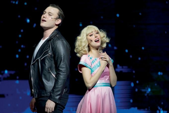 Em roupas do musical Grease, um casal canta em um palco de teatro