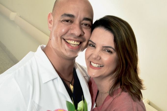 Um casal sorri abraçado e com rostos colados para a foto. Eles estão de roupa de enfermeiros