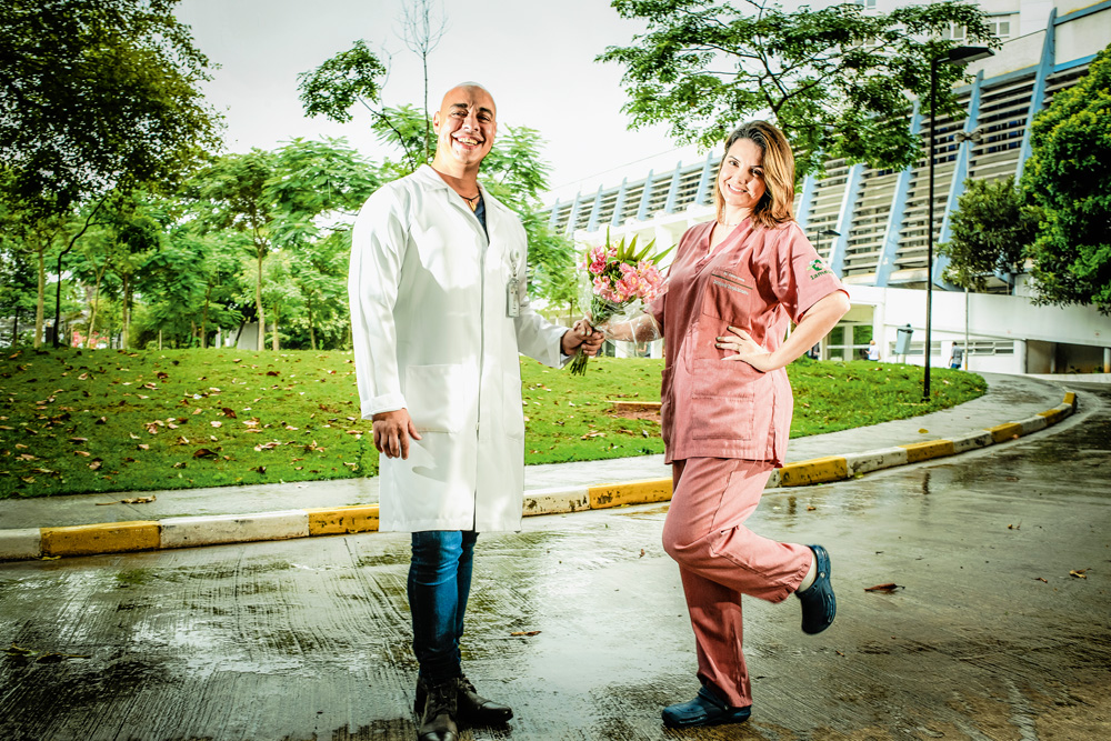 Em frente a um hospital, um casal posa de pé com roupas de enfermagem. Ela segura a mão dele e um buquê de flores une as mãos dos dois