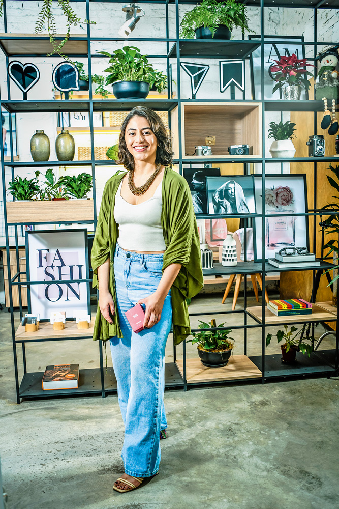 Imagem mostra mulher de calça jeans e top verde em pé, sorrindo para a câmera. Ao fundo, uma estante de metal preta.