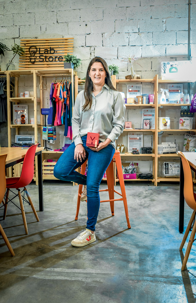 Imagem mostra mulher de calça jeans e camisa cinza com estantes de madeira atrás.