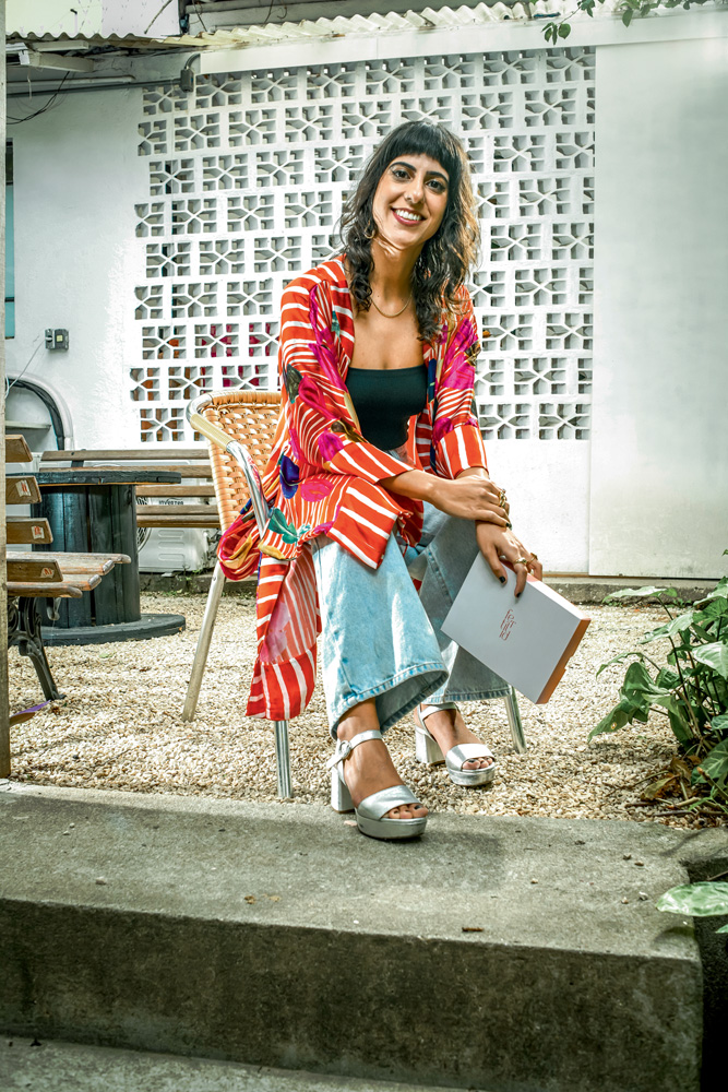 Imagem mostra mulher sentada vestindo roupas coloridas. Ela sorri para a câmera e segura uma caixa branca com uma das mãos.