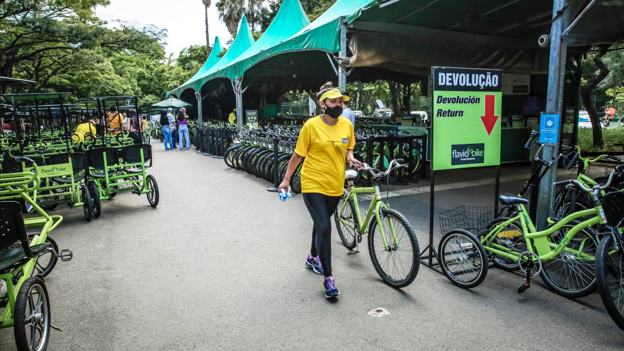 Imagem mostra funcionária da Flavio Bike, vestindo camiseta amarela, levando uma bicicleta. Imagem mostra também local onde são alugados veículos de duas e três rodas