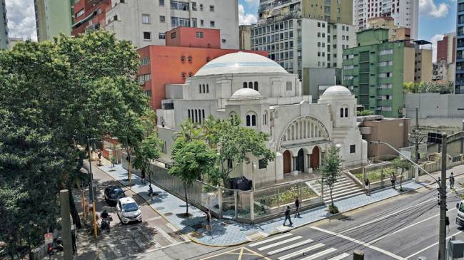 Uma foto um pouco longe de uma antiga sinagoga, com algumas árvores em volta e em um cruzamento