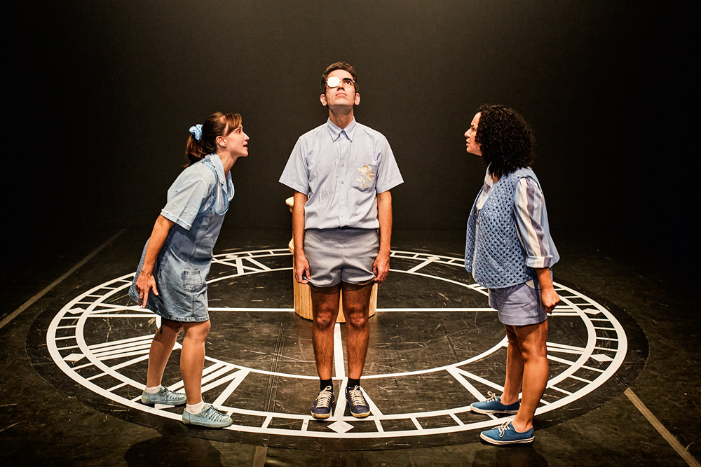 Em palco de teatro, três jovens com roupas azul clara conversam de pé