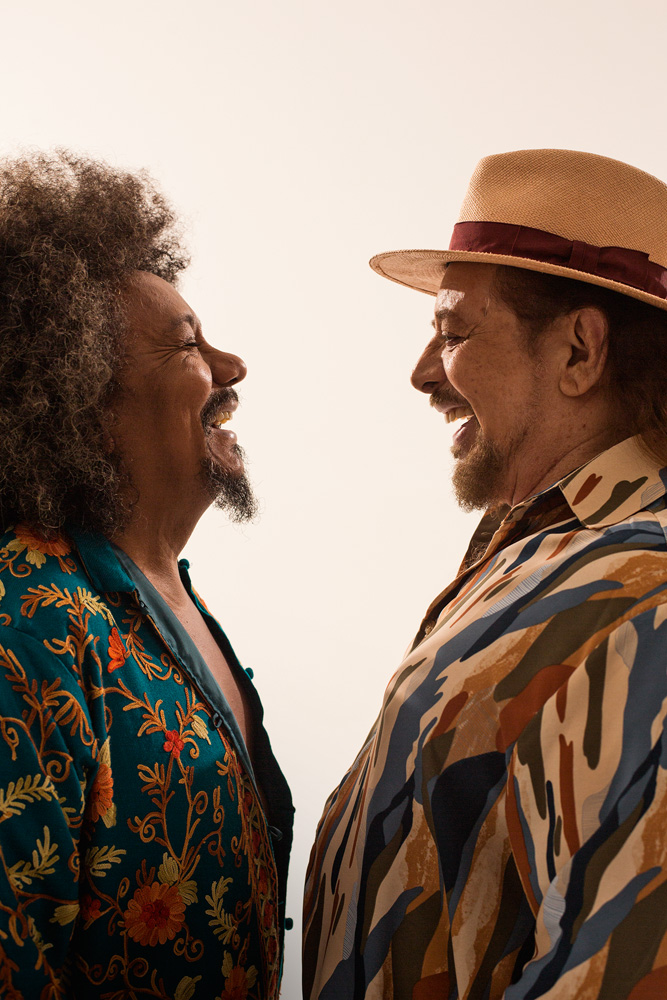 Geraldo Azevedo _ Chico César 2_fotos @marcoshermes.jpg.jpg Imagem mostra dois homens sorrindo, de perfil, se olhando. Os dois vestem camisas estampadas.