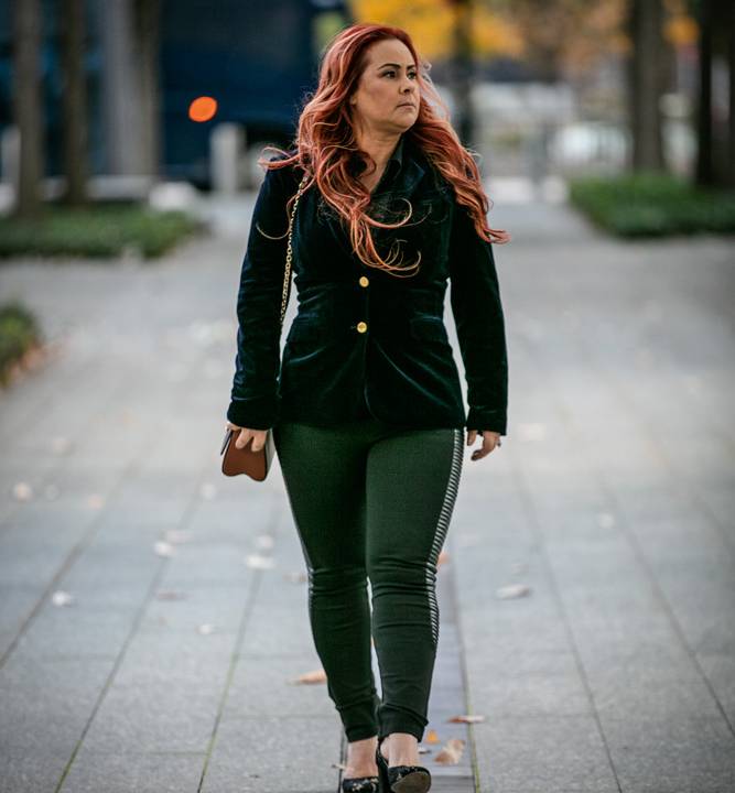 Gracienne olha para a direita enquanto anda em linha reta em grande calçada. O vento bate e leva seu cabelo avermelhado à direita, por cima do blazer preto.