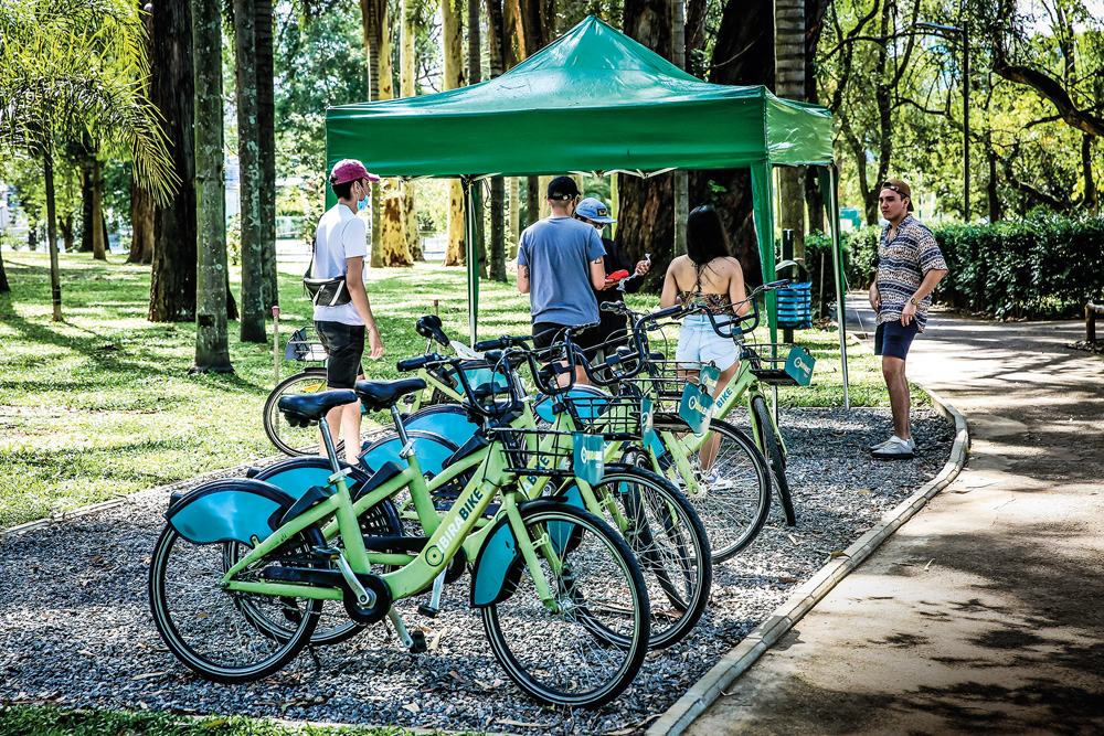 Imagem mostra bicicletas da cor verde claro efileiradas. Ao fundo, árvores e pessoas caminhando.