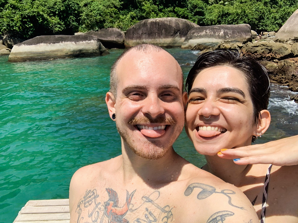 Um casal posa para a selfie em frente a um mar esverdeado em Paraty