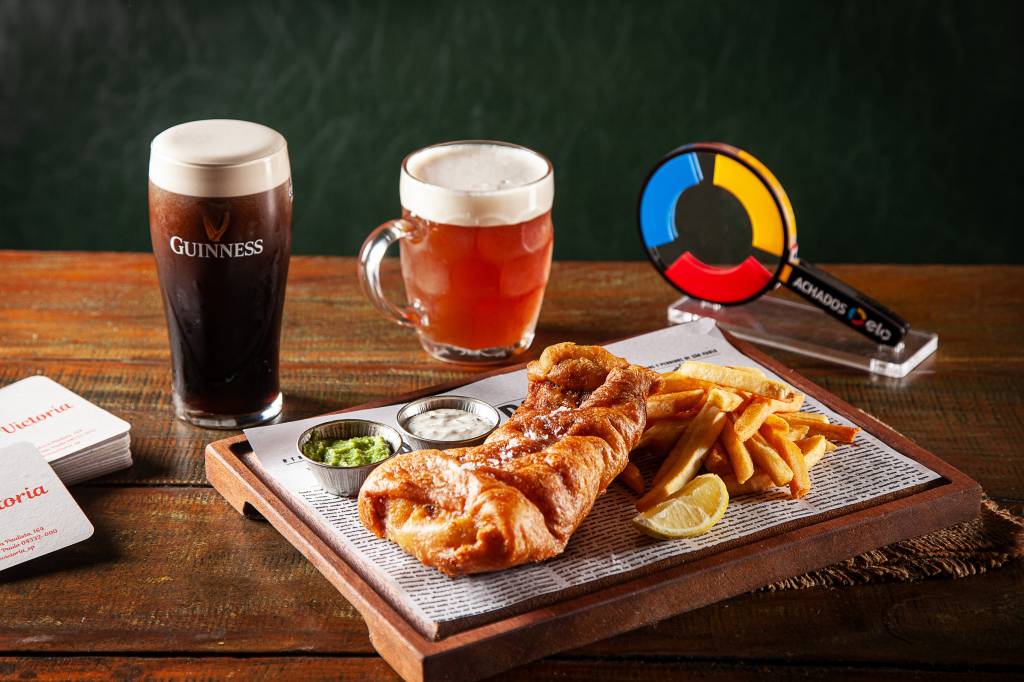 Tábua com fish & chips sobre mesa de madeira ao lado de copo e caneca de chope.