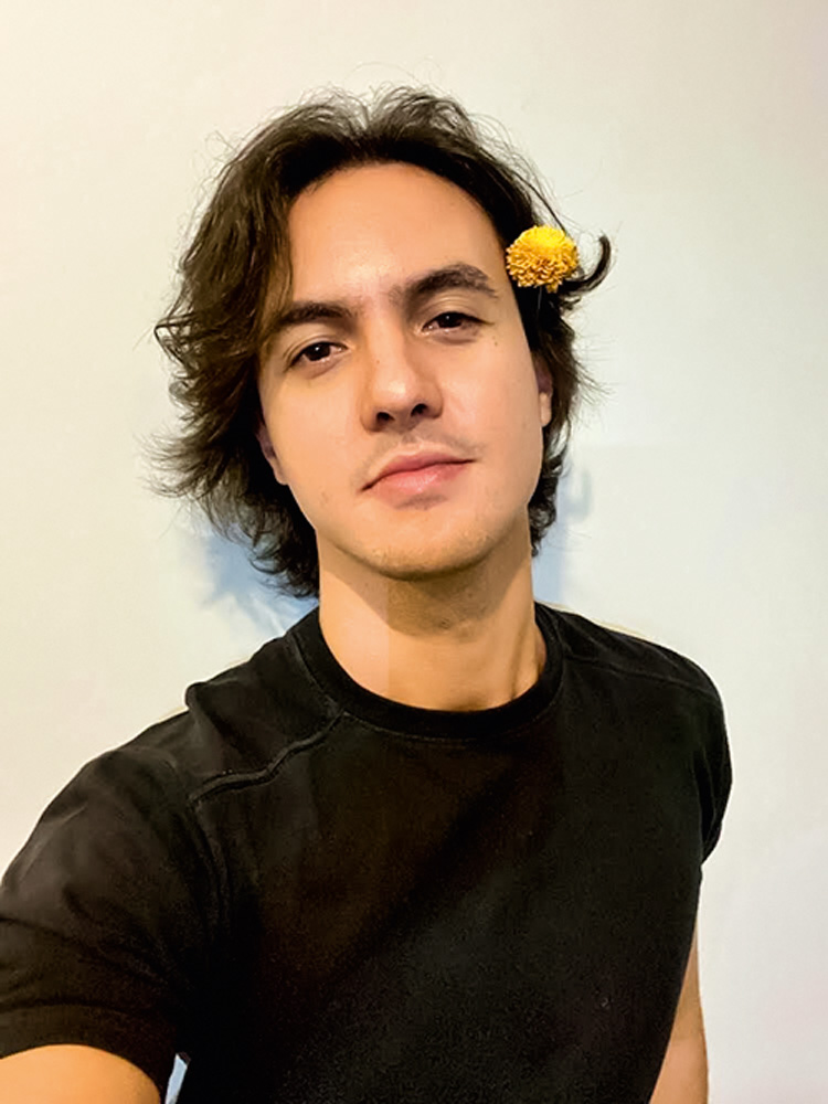 Foto de Renato. Ele é um homem branco, sem barba, de cabelos compridos e escuros. usa uma camiseta preta e uma flor amarela no canto da orelha.