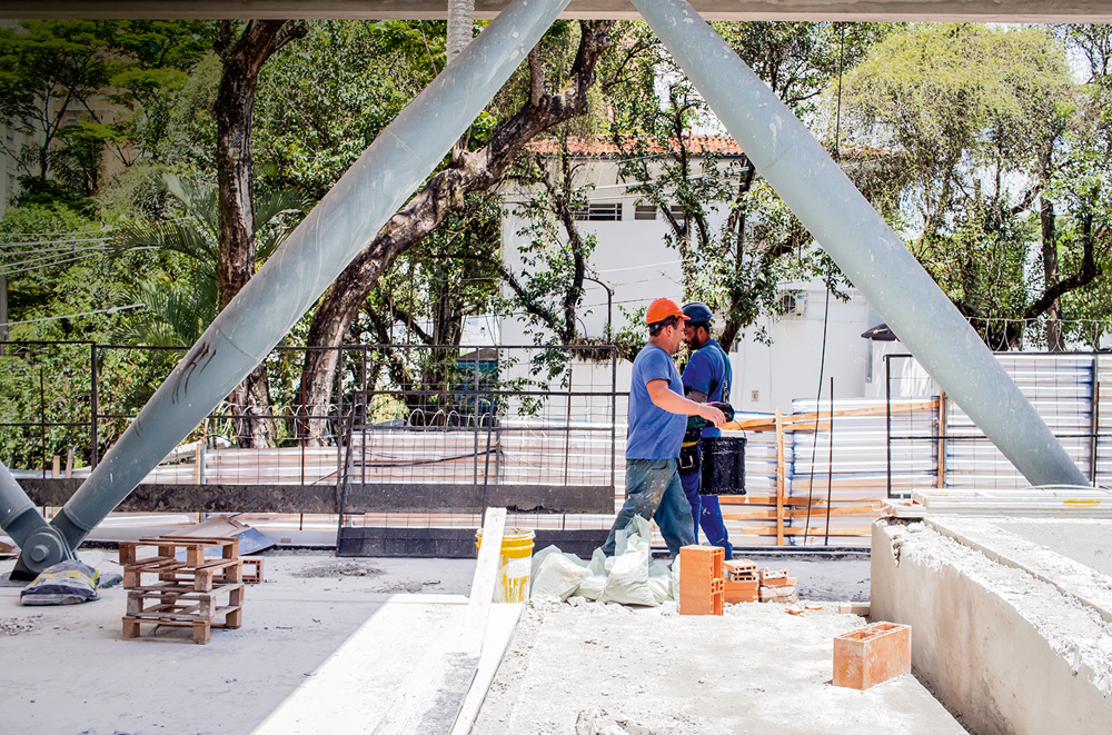 IMG_6916.jpg Imagem mostra dois trabalhadores de capacete e camiseta azul em canteiro de obras com tijolos e cimento fresco.