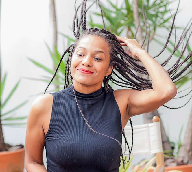 A cineasta Kelly Castilho sorri olhando para baixo enquanto cabelos afro fazem movimento no ar. Ao fundo, plantas e cadeira branca.