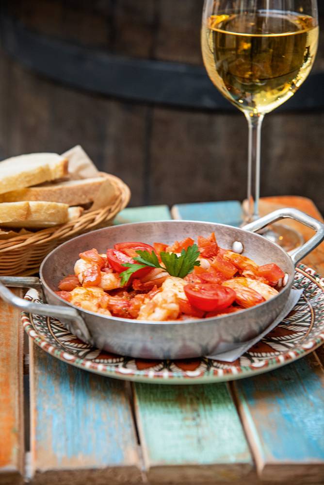 Pequena panela metálica serve camarões com tomate. Ao fundo, uma taça de vinho branco.