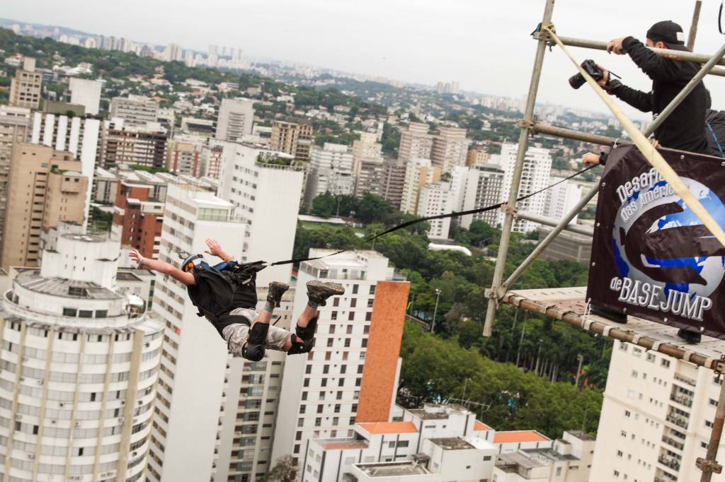 Imagem mostra homem saltando de paraquedas. Abaixo, via movimentada do centro de São Paulo