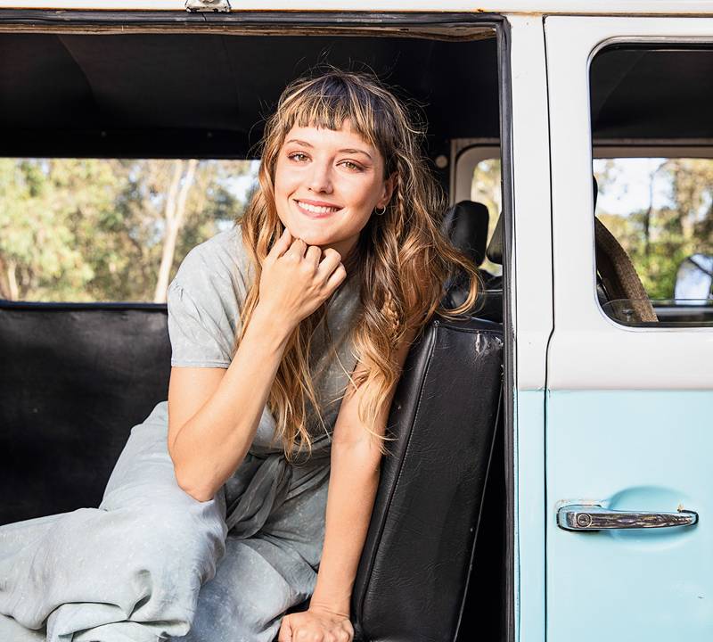 Imagem mostra mulher de vestido azul claro sorrindo sentada no banco de uma Kombi branca e azul.