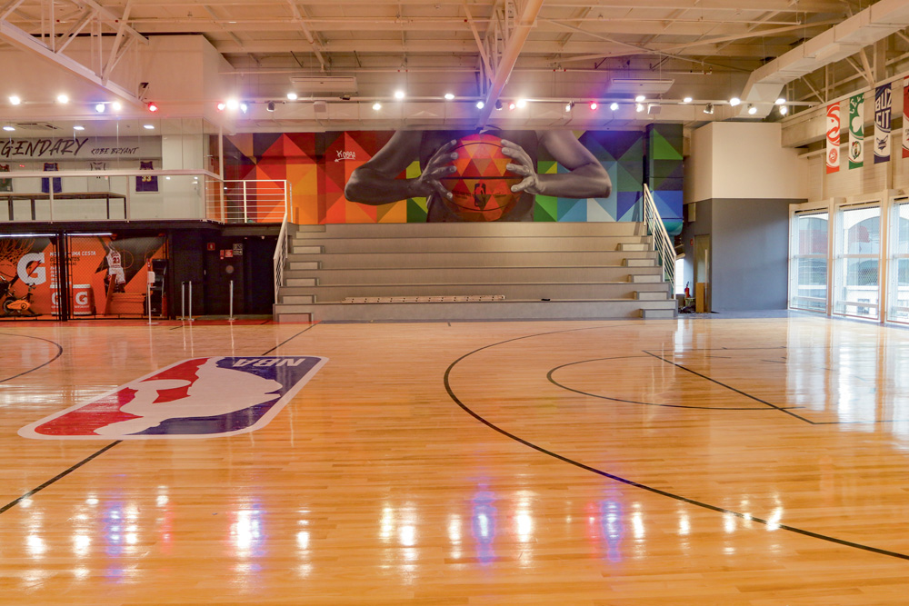 Foto mostra uma quadra de basquete, com arquibancada e artes ao fundo e o símbolo da NBA na quadra