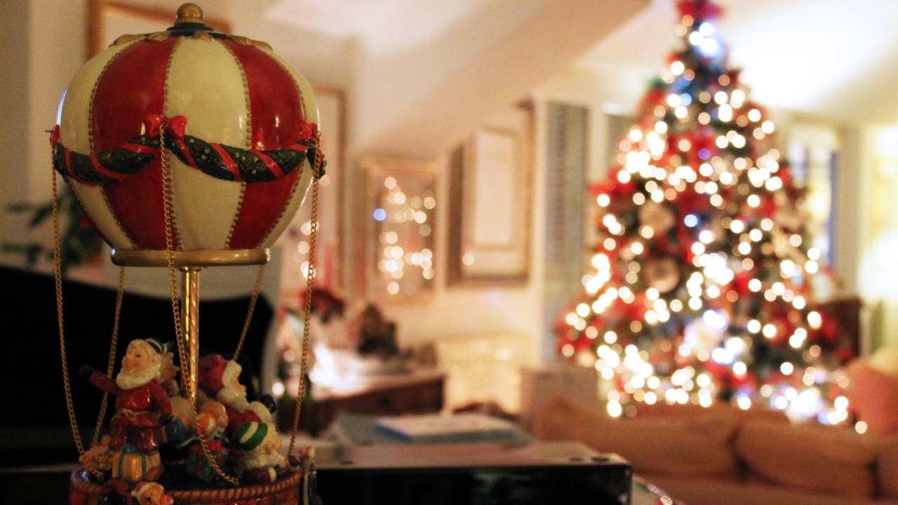 Foto exibe sala decorada com objetos de Natal. Em primeiro plano, um balão branco e vermelho de cerâmica. Ao fundo, uma grande árvore natalina com luzes brancas.