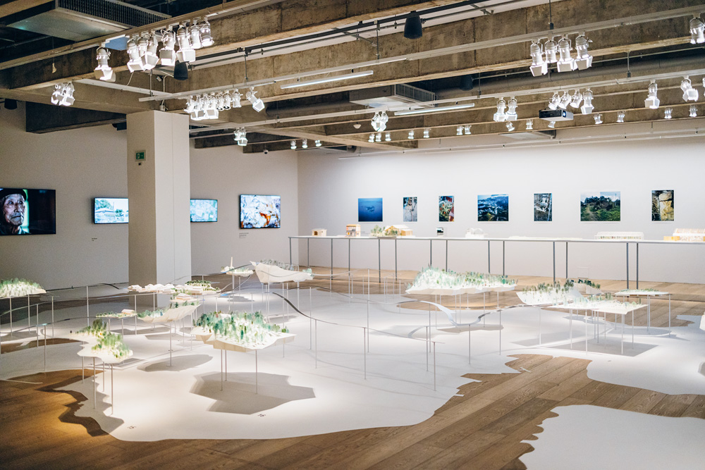 Imagem mostra a exposição em sala da Japan House. O chão é de madeira e as paredes são brancas. É possível ver várias maquetes distribuídas pelo espaço e televisores que exibem vídeos nas paredes.