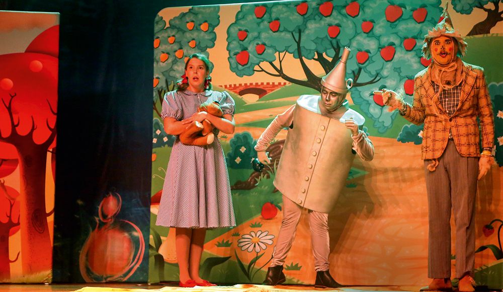 Em um palco de teatro todo decorado, atores vestidos de Dorothy, Espantalho e Homem de Lata interpretam O Mágico de Oz