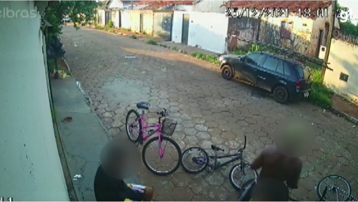 Imagem mostra uma rua com dois meninos na calçada, com duas bicicletas no chão.