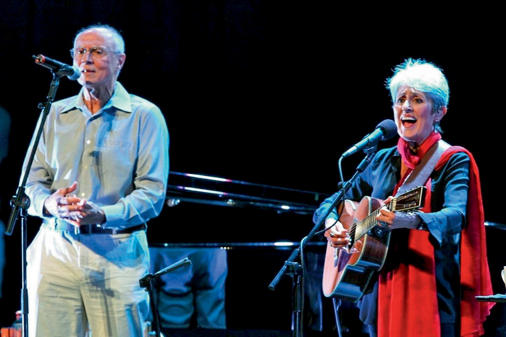 Suplicy com Joan Baez cantando no palco. Ela toca um violão e usa roupa vermelha. Suplicy está ao lado e usa uma camisa vermelha e uma calça branca. Ambos têm cabelos curtos e grisalhos.