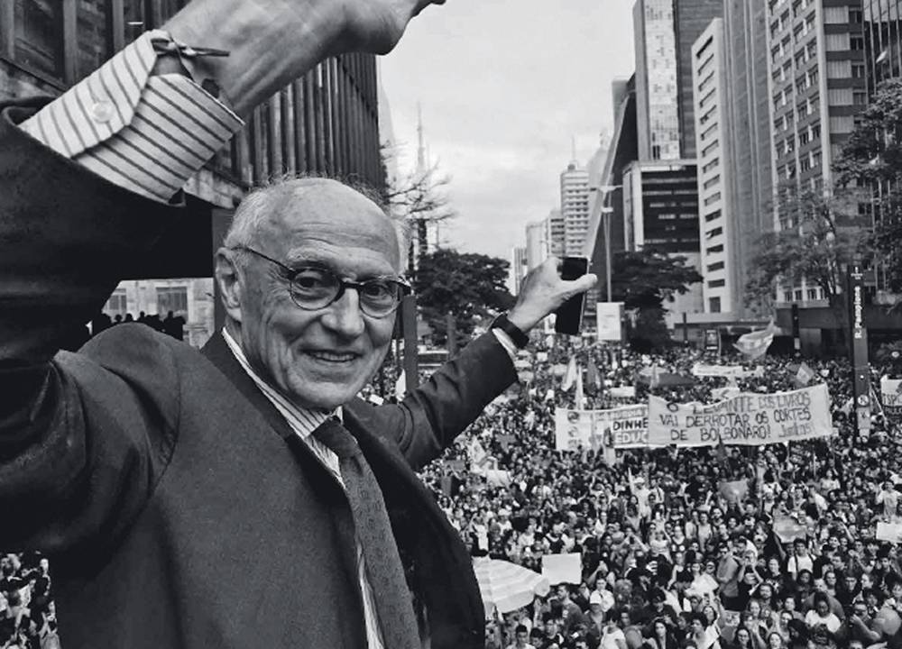 Foto em preto e branco do ex-senador Eduardo Suplicy em manifestação na Avenida Paulista. Ele aparece de terno, óculos e levanta os braços para cima em comemoração.