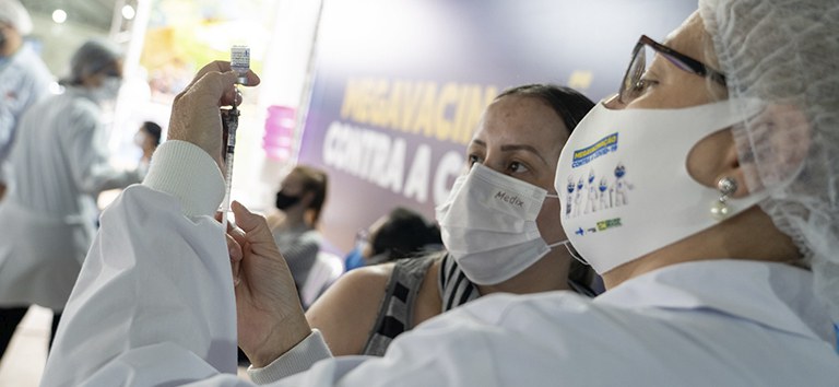 Profissional de saúde vestida de branco segurança seringa e vacina contra a Covid-19 e mostra para uma outra mulher