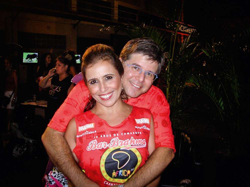 Imagem mostra um homem abraçando uma mulher por trás. Os dois vestem camisetas vermelhas.