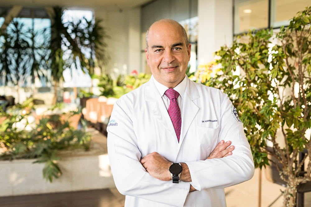 A imagem mostra Fernando Ganem, diretor-geral do Hospital Sírio-Libanês, posando para uma foto. Ele é um homem branco, calvo e de cabelos grisalhos. Veste um jaleco branco com seu nome e uma gravata vermelha.