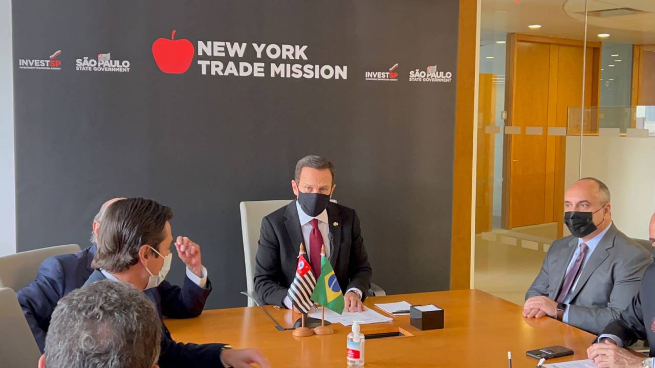 Imagem mostra, na cabeceira da mesa, João Doria sentado usando terno e gravada. À frente, bandeirinhas do Brasil e de São Paulo. Na parede ao fundo lê-se: "New York Trade Mission", com bandeira de São Paulo e logotipo da InvestSP