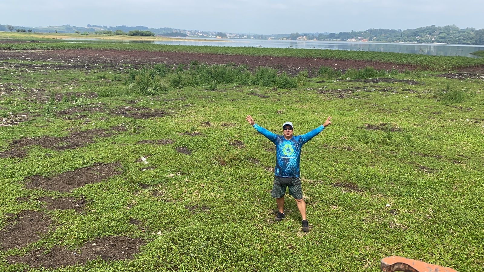 Imagem mostra homem vestido de camiseta azul abrindo os braços em meio a gramado da represa de Guarapiranga