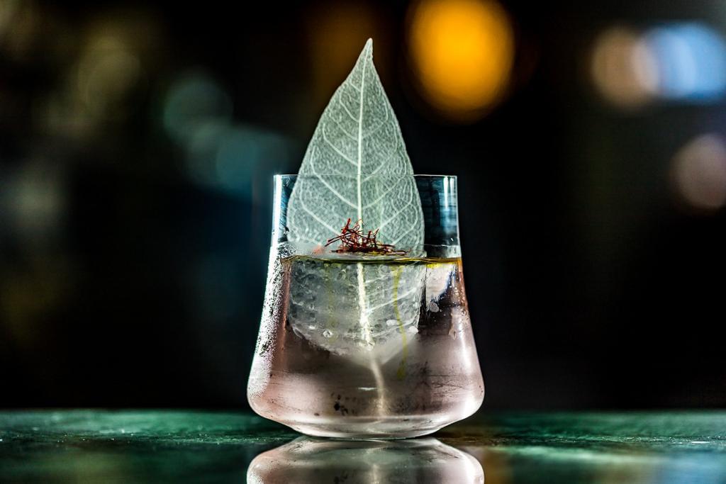 Drinque quase transparente servido em um pequeno copo de fundo redondo decorado por uma folha verde
