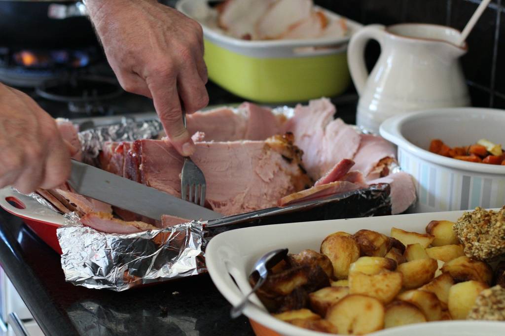 Imagem mostra mesa cheia de comida e mãos cortando pedaço de carne de porco com garfo e faca