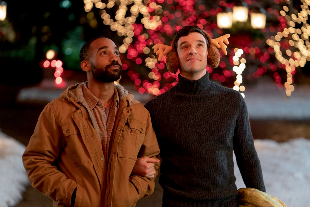 Imagem mostra dois homens, um com um casaco marrom e um com um suéter preto. Ao fundo, luzes de natal.