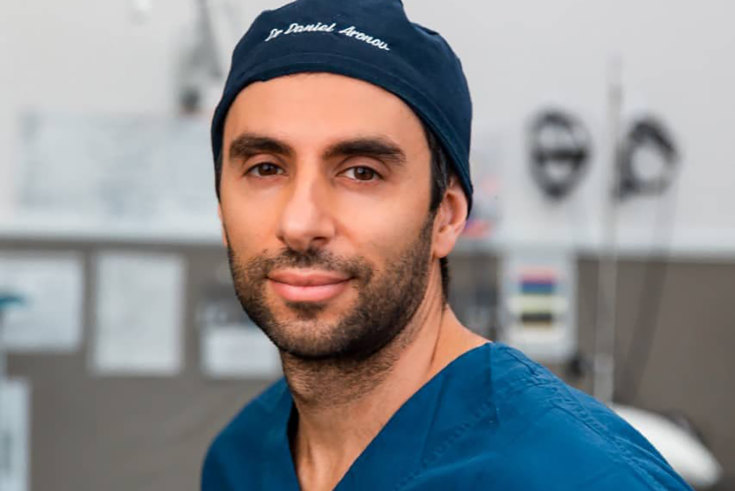 Imagem mostra Daniel com touca em que seu nome está bordado; ele usa avental médico e, ao fundo, sala de cirurgia