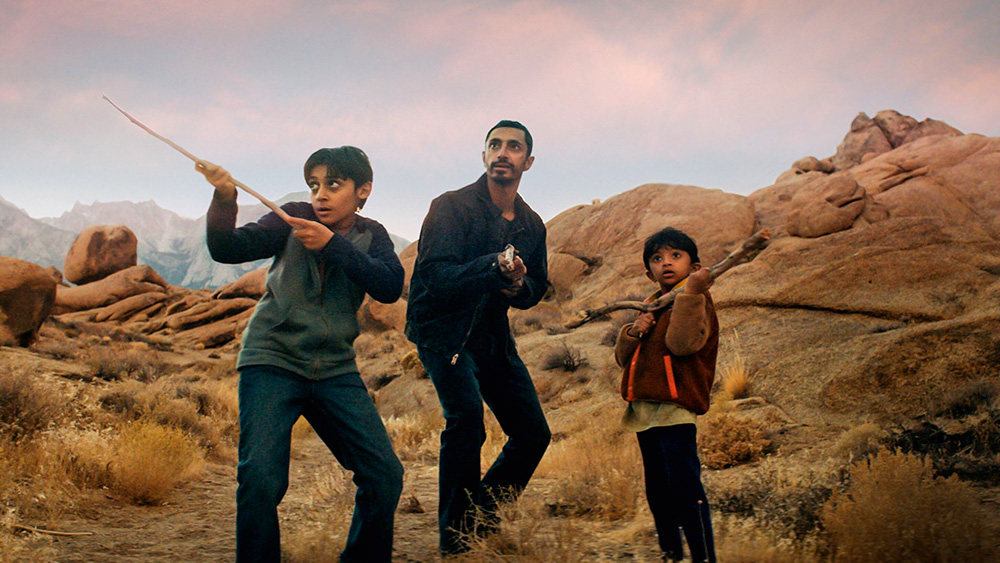 Imagem mostra um homem e dois garotos em um deserto. Os garotos seguram gravetos, o homem segura um rifle.
