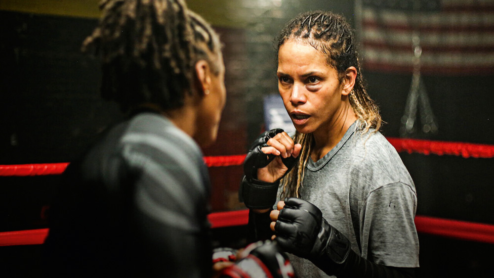 Imagem mostra duas mulheres, uma de costas e uma de frente para a câmera. Elas vestem luvas de boxe e estão em um ringue.