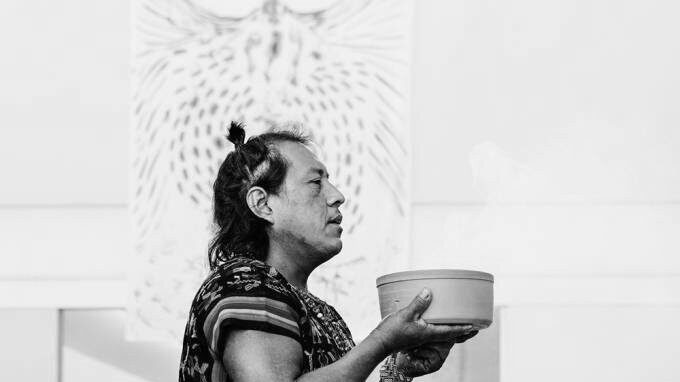 Artista Jaider Esbell com um vaso nas mãos e parte dos cabelos presos