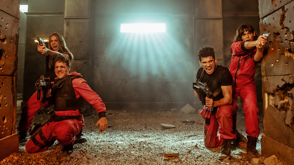 Imagem mostra 4 pessoas de macacões vermelhos e coletes à prova de balas segurando armas. Elas estão em um local repleto de escombros, atrás de pilastras com marcas de tiro.