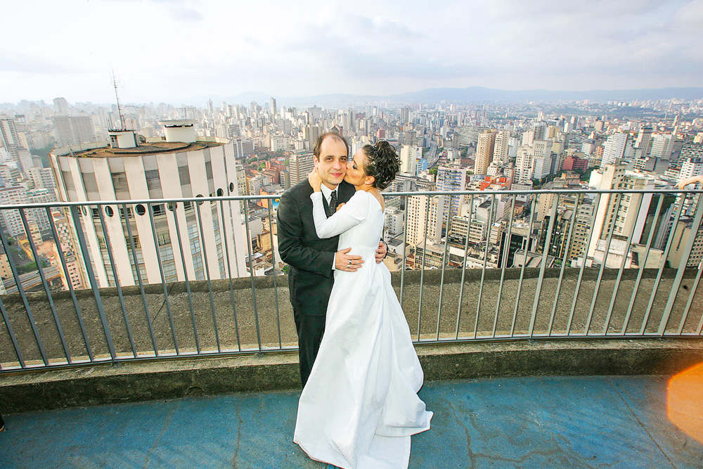 Imagem mostra um homem e uma mulher, ele de terno e ela de vestido branco, em terraço de prédio. Ao fundo, um horizonte de prédios.