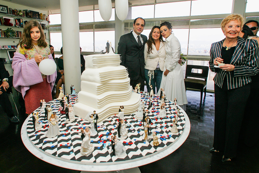 Imagem mostra uma mesa com um bolo branco em formato ondulado. Ao redor da mesa, uma menina, uma mulher e um casal com terno e vestido branco.