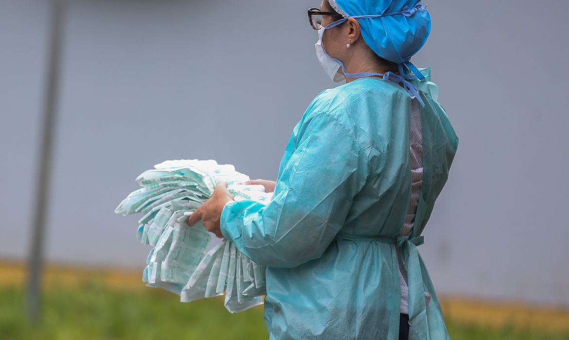 Uma profissional da saúde segura seringas embaladas em plástico. Ela veste um traje azul, máscara, touca e óculos.
