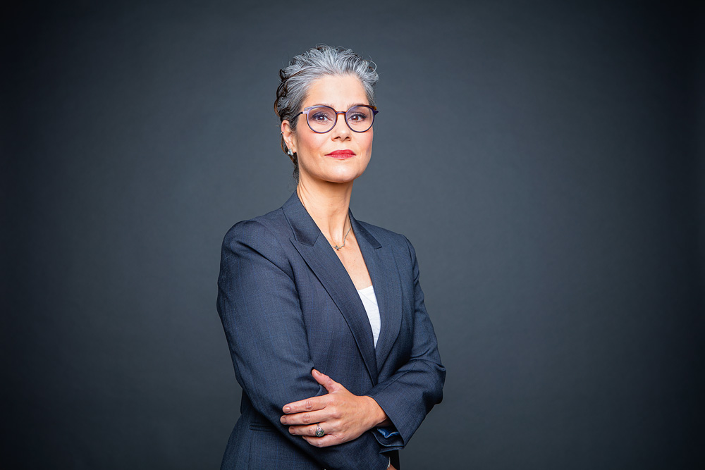 Patricia Vanzolini aparece posando para a foto. Ela é focalizada da cintura para cima, veste uma blusa branca e um blazer cinza, que combina com o fundo também na cor cinza. Ela é uma mulher branca, de cabelos grisalhos e usa óculos.