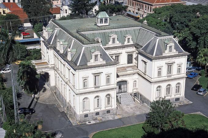 Imagem mostra construção clássica branca com telhado preto. Prédio de dois andares com paredes barroca.