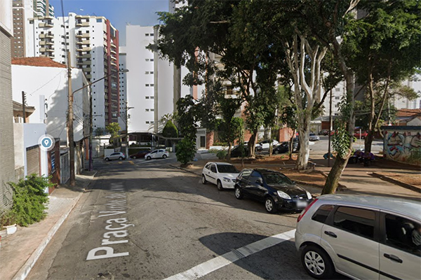 Imagem mostra rua com praça arborizada na lateral direita. Ao fundo, prédios residenciais