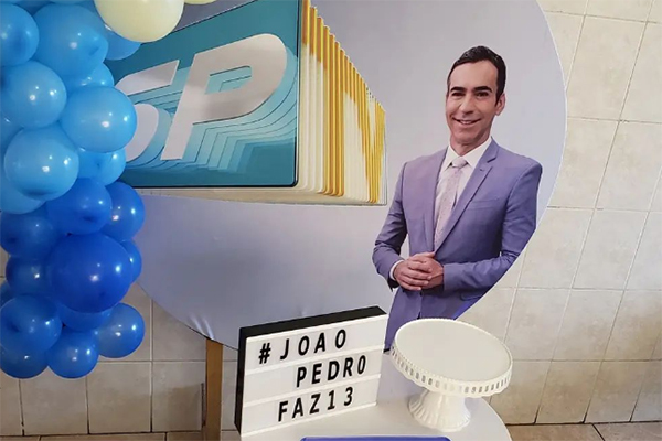 Imagem mostra mesa de aniversário, com balões, painel que diz "#João Pedro faz 13" e foto de César Tralli, ao lado de logo do SPTV, ao fundo