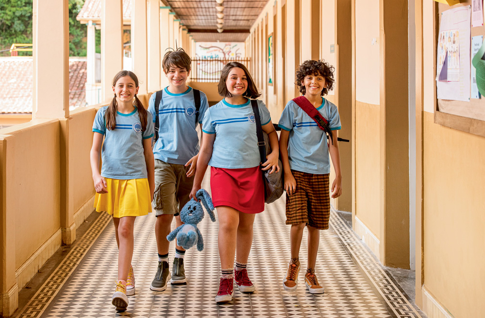 Imagem mostra 4 crianças com uniforme azul claro andando em corredor.