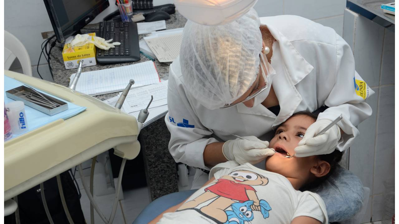 Imagem mostra criança em consultório de dentista, sendo atendida por uma dentista.